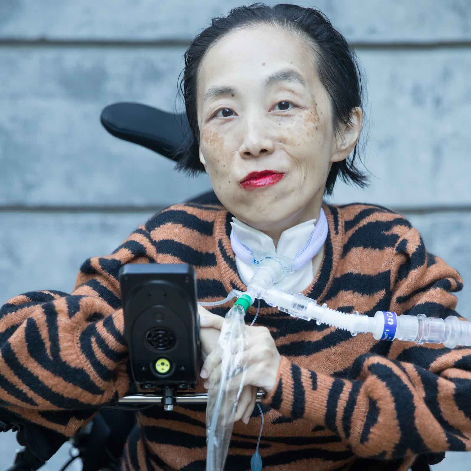 Alice Wong, an Asian American disabled woman in a power chair. She is wearing a black blouse with a floral print, a bold red lip color and a trach at her neck