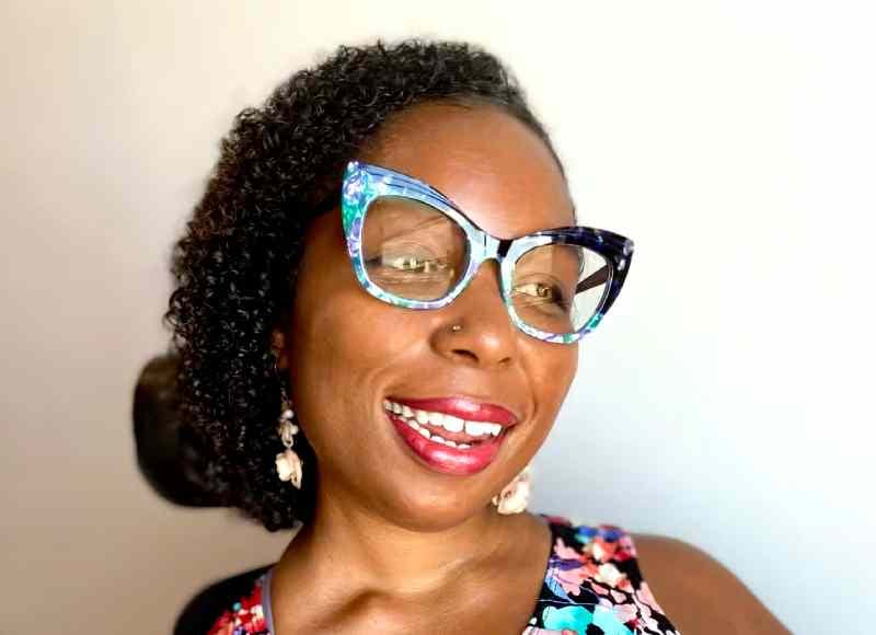 Andraéa LaVant flashes a large smile as she shows a three-quarters profile looking past the camera. Her head and shoulders are visible in the photo. She is wearing a colorful flower print dress, red lipstick, and fun, colorful glasses.