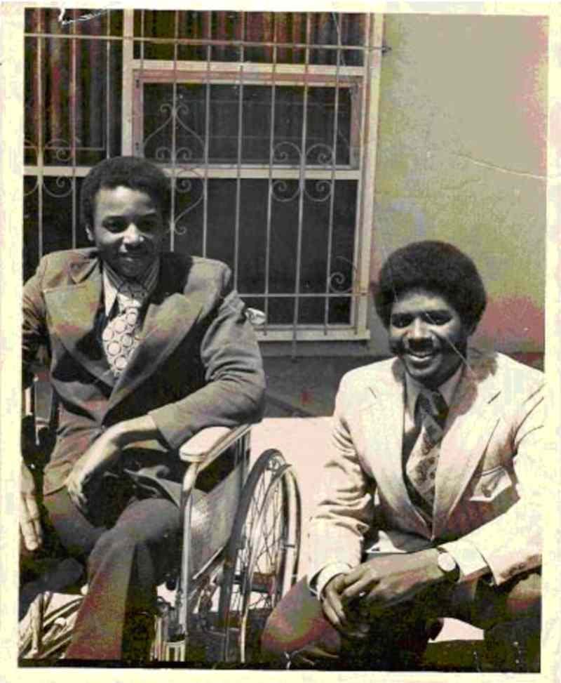 Brad and Glenn Lomax smile for the camera wearing suits in this black and white photo. Brad is seated in a wheelchair. Glenn squats beside him.