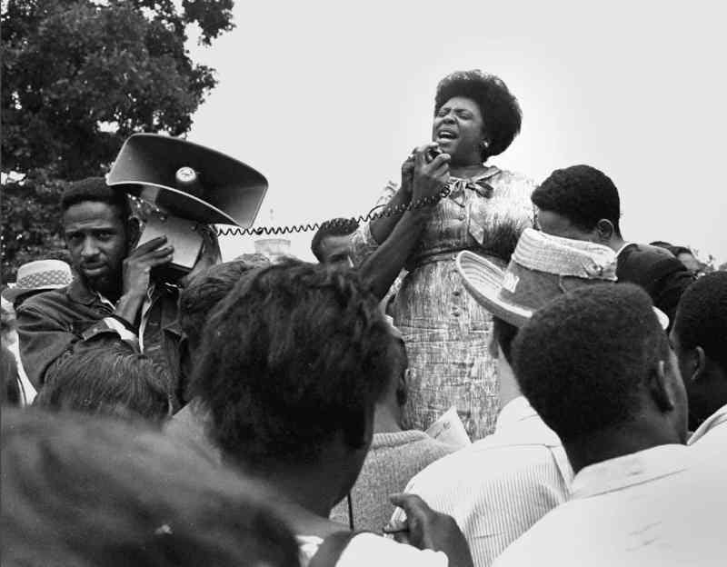 Fannie Lou Hamer stands on a raised platform surrounded by people holding the microphone for a loudspeaker being held up by a man a couple of feet away from her. She has a determined and passionate look on her face as she talks with the assemebled crowd.