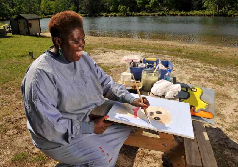 Lois Curtis sits at a small picnic table next to a lake with her art supplies as she paints a portrait of a dog. She has a big smile on her face and her frock is covered with drops of paint.
