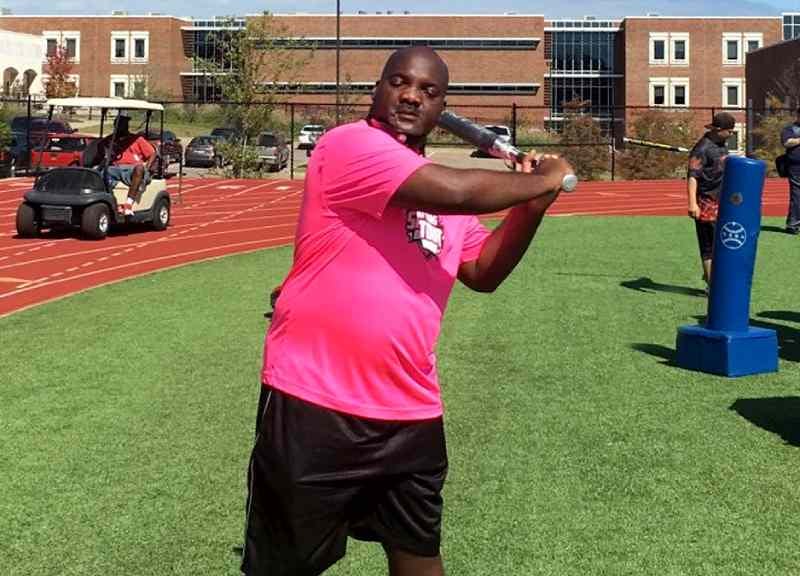 Tracey Jackson poses with a bat at a Beep Baseball event. He is wearing black shorts and a bright pink T-shirt and is standing on a school sports field.