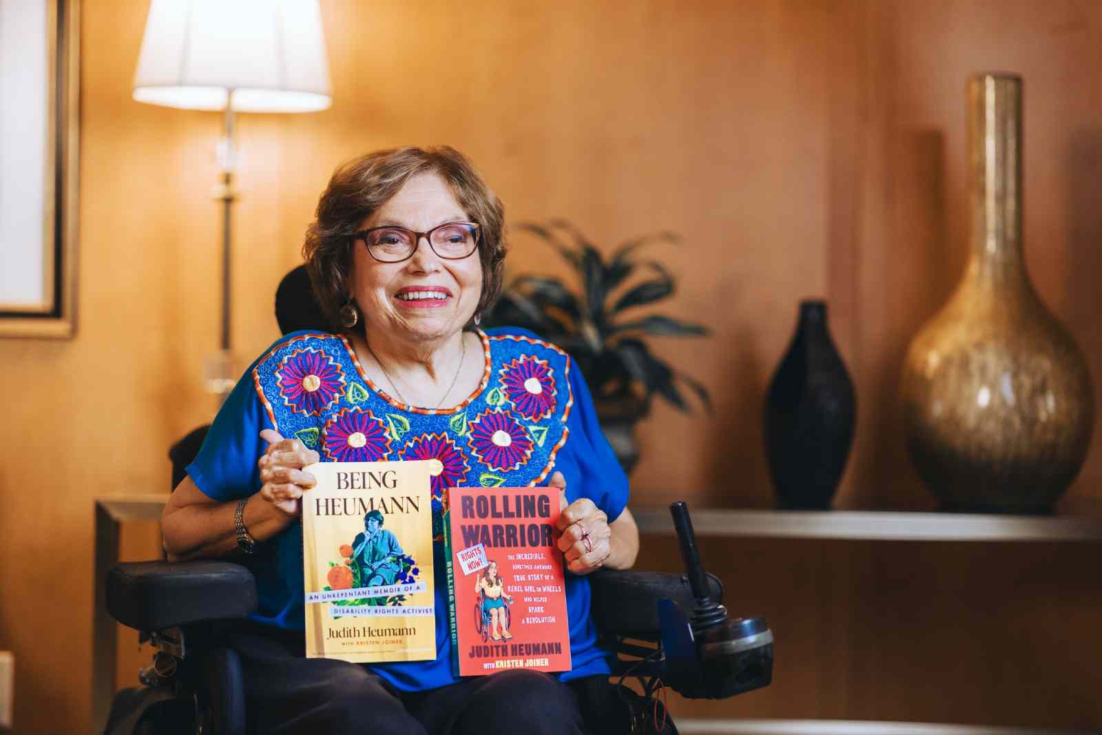 Judy Heumann in her wheelchair holding her two seminal books - “Being Heumann” and “Rolling Warrior”