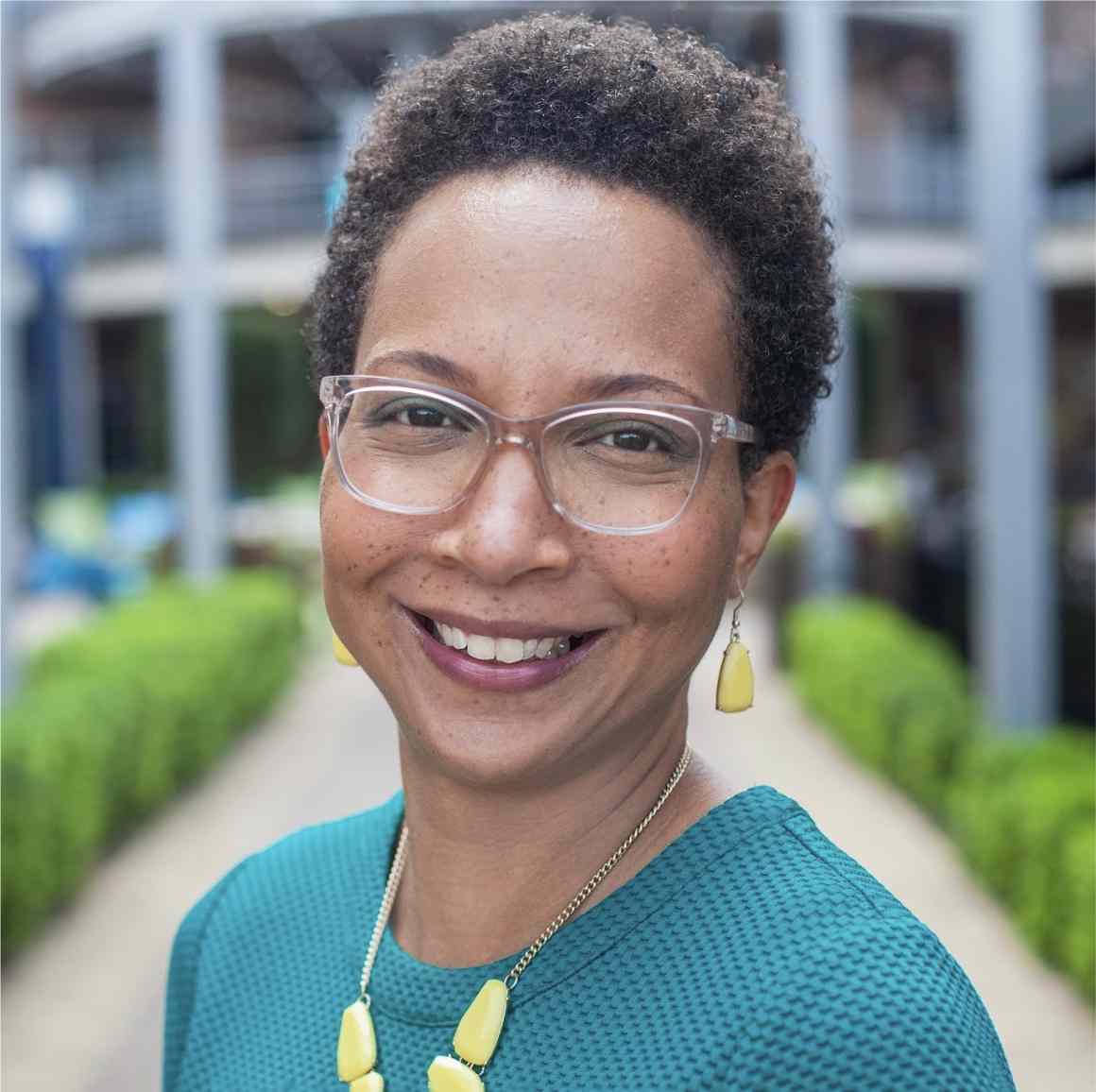 Outdoor professional headshot of Dr. Michele Williams. She is a Black woman pictured with short natural hair and clear rimmed eyeglasses.